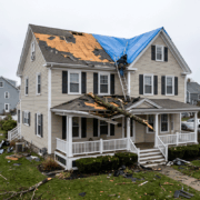 Two-story beige house with a torn roof covered by a blue tarp, a worker on a ladder, and debris scattered across the yard after a storm fall.