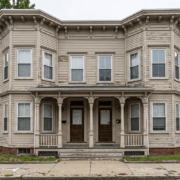 Two-story beige wooden house with peeling siding and a centered front porch supported by decorative columns, two front doors side by side.