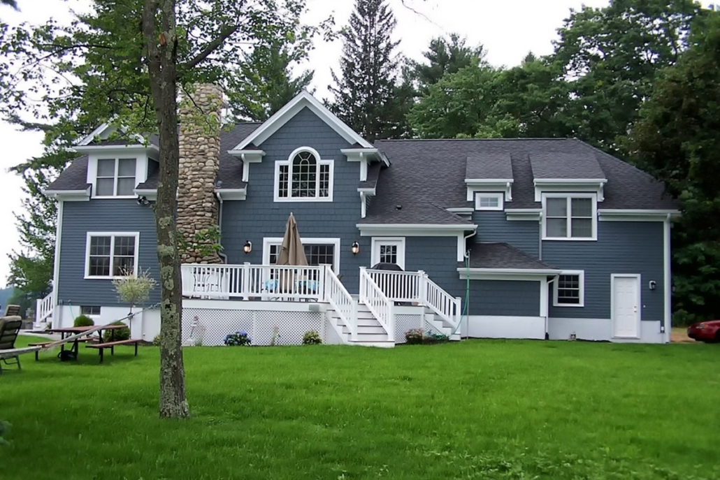 Two-story blue house with white trim, a stone chimney, and a raised white deck overlooking a green lawn with trees nearby. | J.E.P. Contracting