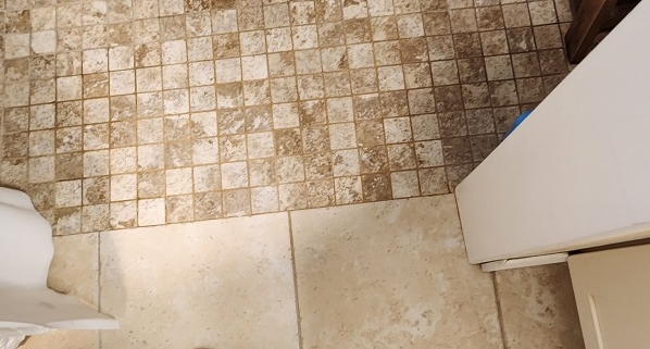 Top-down view of a person standing on a beige bath mat in a tiled bathroom, wearing gray sneakers and light shorts beside a wooden stool.