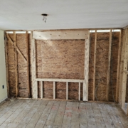 Room under renovation with exposed wooden studs, OSB wall sheathing, and a ceiling electrical hole with hanging wires.